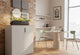 Moda Office Storage Cabinet in a collaborative office setting with exposed brick and white walls, high ceilings, tall windows, and a table with two chairs. The cabinet features a durable powder-coated surface, ABS handles, damping buffer hinges, a front lock, and a laminate top, providing functional and stylish storage for modern workspaces.
