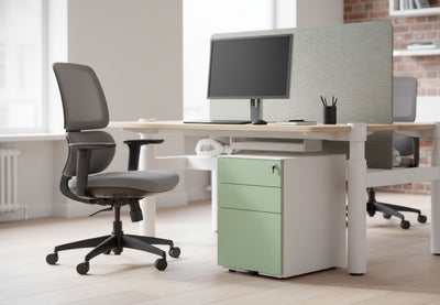 Moda Mobile Filing Cabinet positioned under a desk in an office-style interior with exposed brick walls and wooden flooring, showcasing its powder-coated steel construction, smooth Hettich slide rails, secure front lock and POM casters; modern mobile office storage designed for efficient, space-saving organisation.