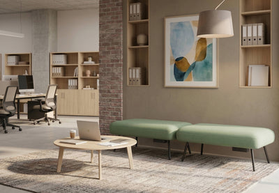 Two Omni Upholstered Office Benches are arranged on a patterned rug, creating a flexible touchdown point in a modern open-plan office. Each green upholstered bench, made with durable 100% polyester OTE fabric, features a built-in plug socket for convenient device charging of laptops and mobile devices. A round coffee table with a laptop completes this inviting collaborative zone, designed for flexible working, informal meetings and casual collaboration.