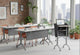 Swish Adaptive Presentation Podium in white oak and black shown in an interior image, positioned at the front of a classroom-style space with four tables facing it, surrounded by high ceilings, large windows and exposed brick walls; modern mobile presentation podium designed for professional training, teaching and collaborative office environments.
